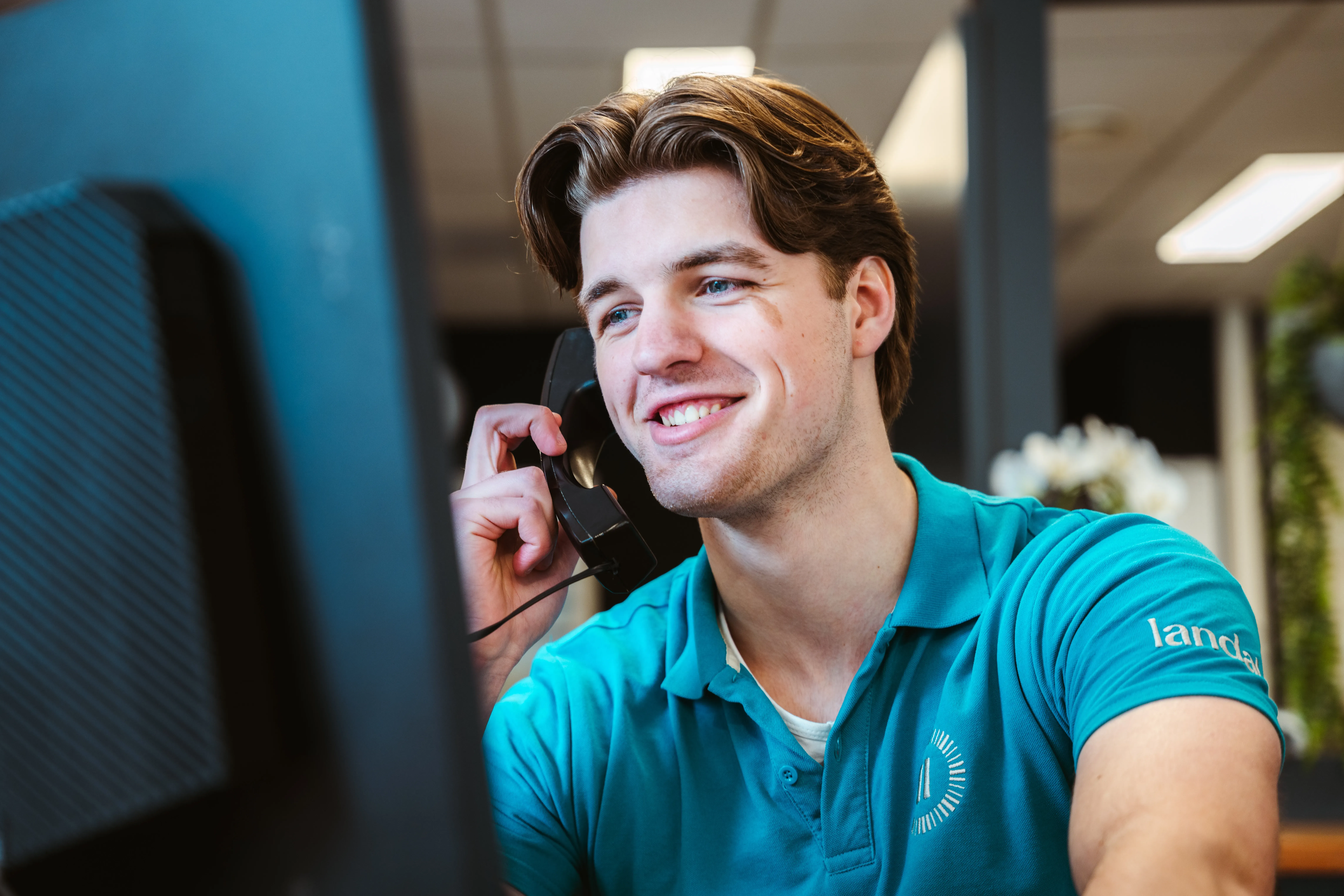 Ein lächelnder junger Landal-Mitarbeiter im türkisfarbenen Poloshirt hält einen Telefonhörer und schaut auf einen Bildschirm in einer hellen Büroumgebung mit Pflanzen im Hintergrund.