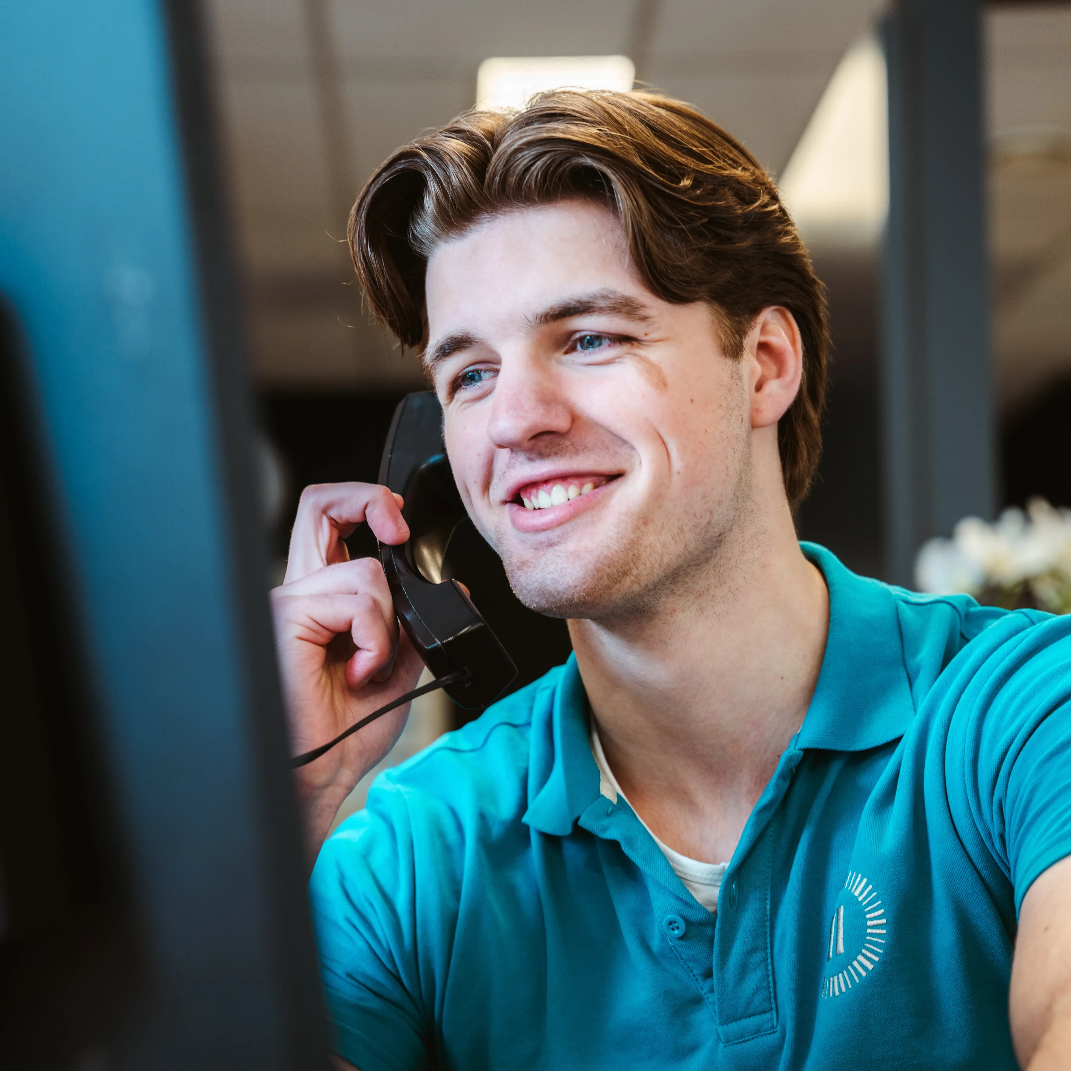 Ein lächelnder junger Landal-Mitarbeiter im türkisfarbenen Poloshirt hält einen Telefonhörer und schaut auf einen Bildschirm in einer hellen Büroumgebung mit Pflanzen im Hintergrund.