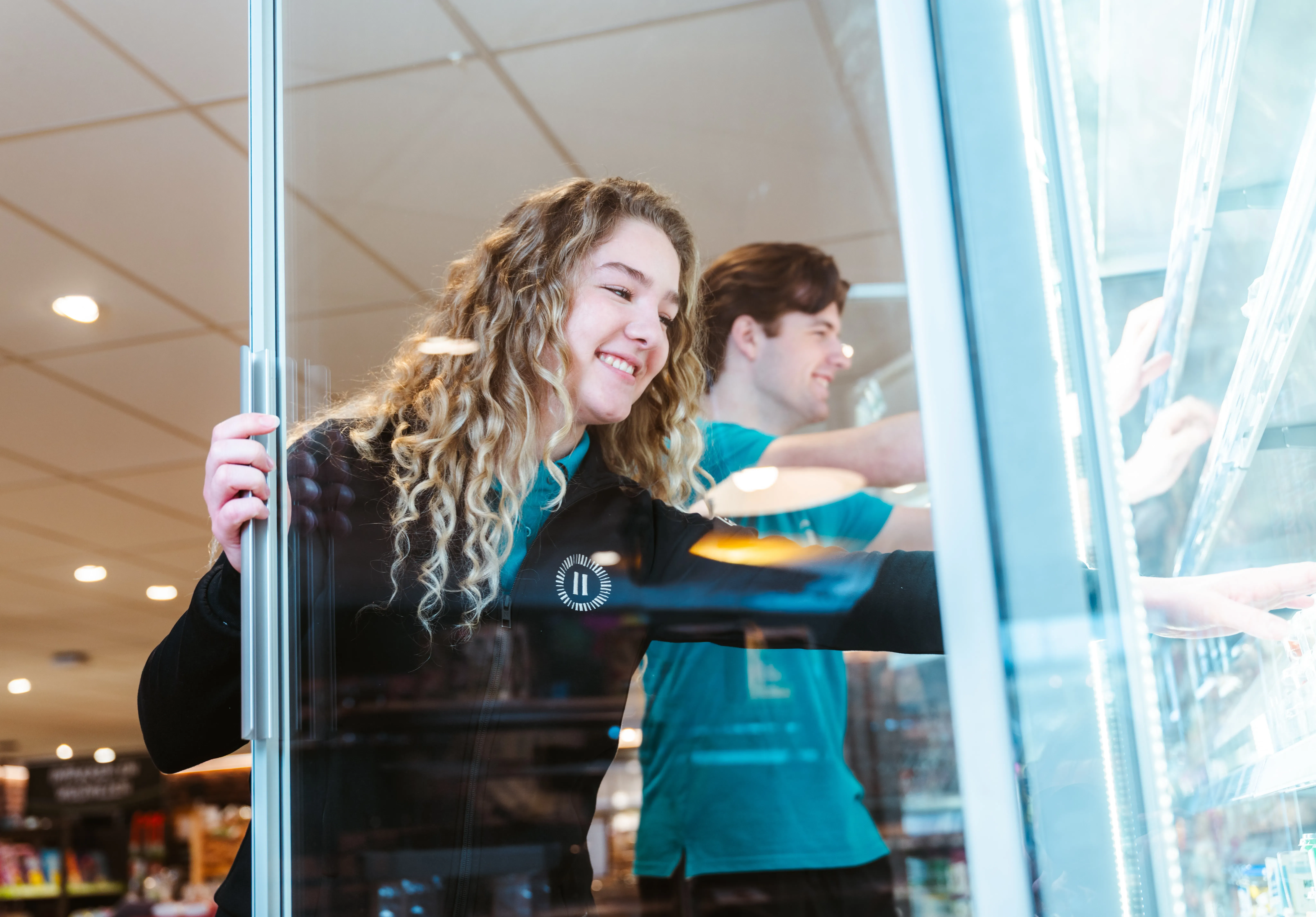 Twee jonge Landal-medewerkers vullen lachend een koelkast bij in een parkshop — zij in zwart uniform op de voorgrond, hij in teal polo op de achtergrond.