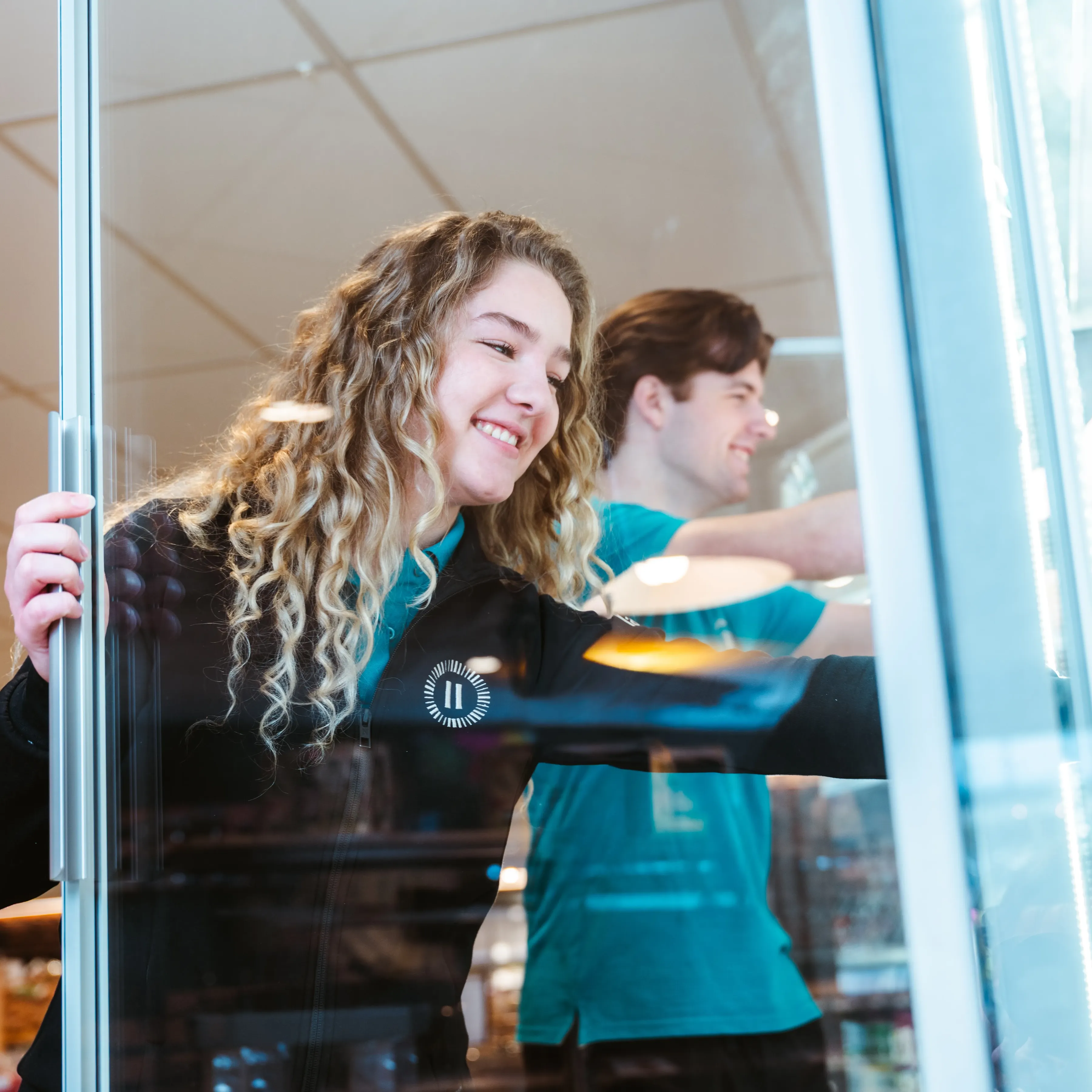 Twee jonge Landal-medewerkers vullen lachend een koelkast bij in een parkshop — zij in zwart uniform op de voorgrond, hij in teal polo op de achtergrond.