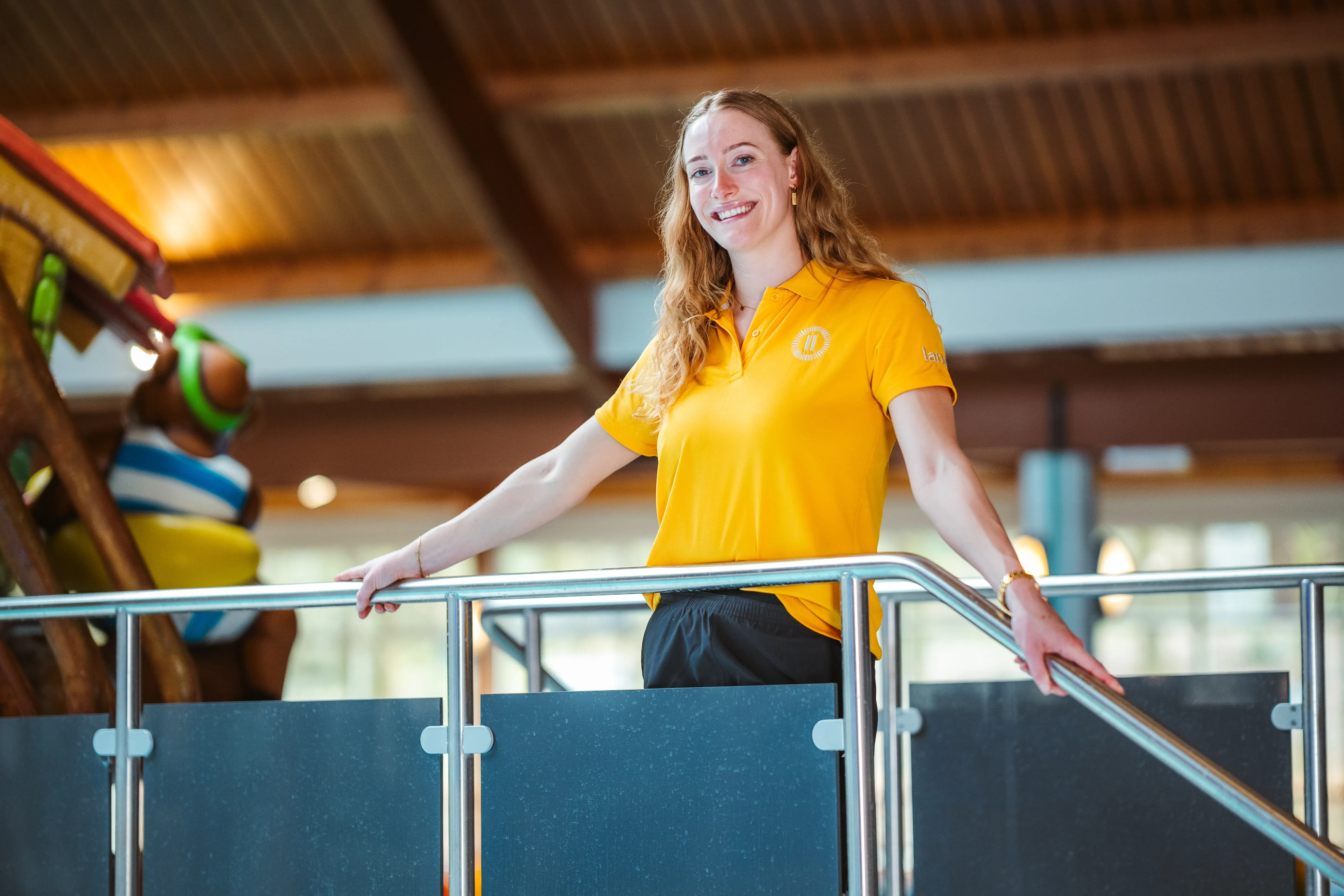 Een lachende jonge Landal-medewerker in geel polo leunt ontspannen op een reling bij het overdekte zwembad, met op de achtergrond een glijbaan en speelattributen.