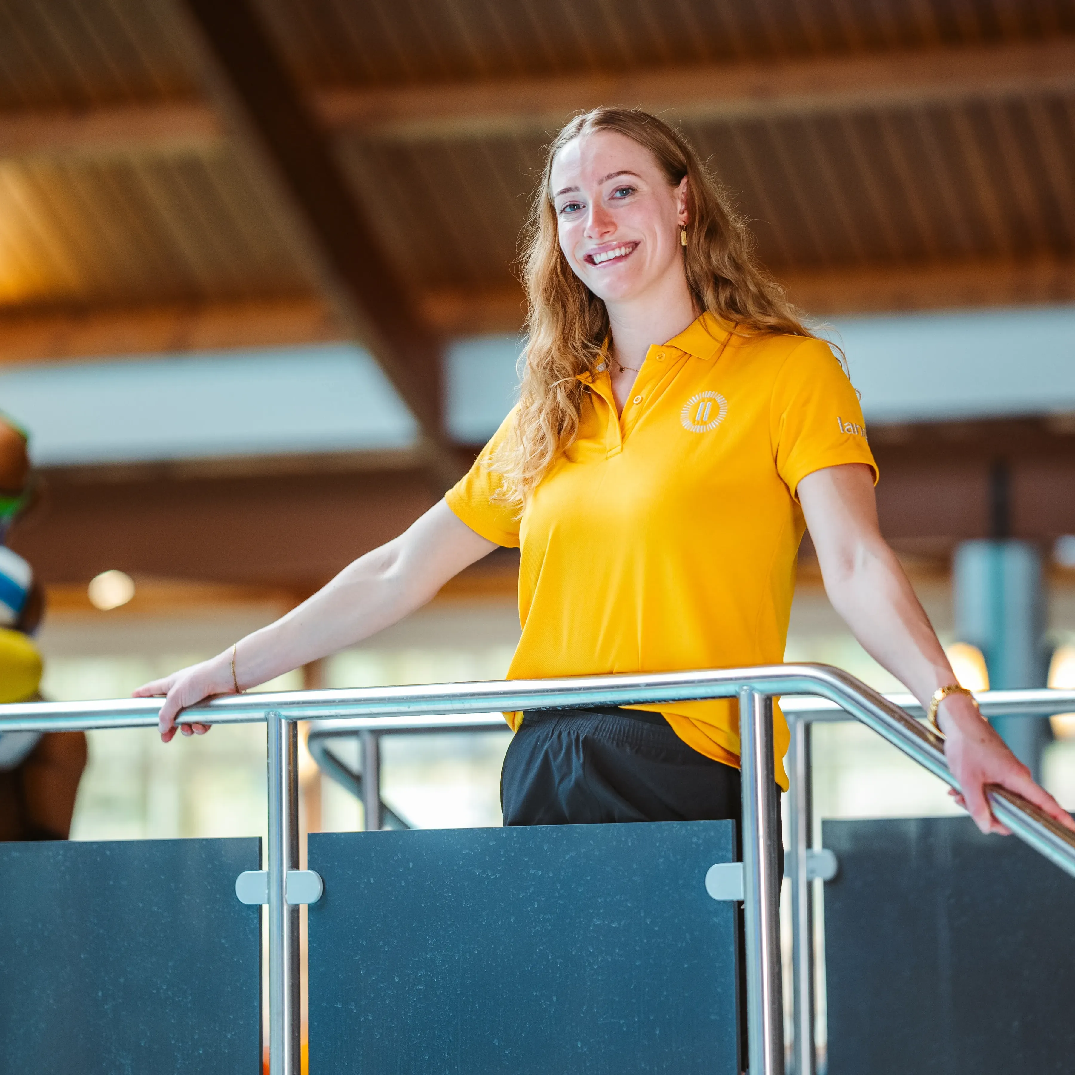 Een lachende jonge Landal-medewerker in geel polo leunt ontspannen op een reling bij het overdekte zwembad, met op de achtergrond een glijbaan en speelattributen.