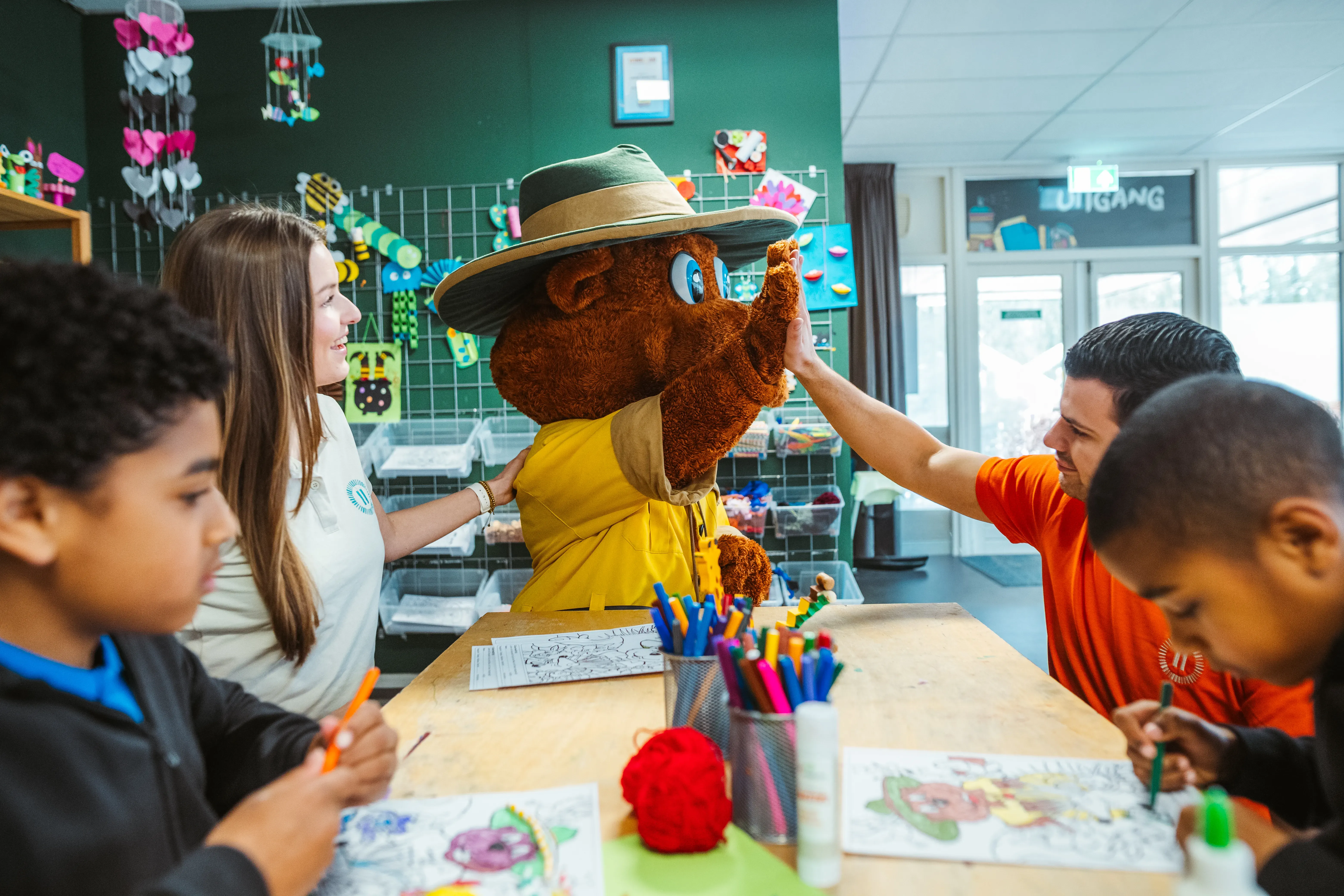 Zwei kleine Jungen sitzen an einem Tisch und malen ein Bollo-Malbild aus. Bollo steht am Ende des Tisches und gibt einem männlichen Landal-Mitarbeiter ein High-Five, während eine Mitarbeiterin Bollo die Hand auf die Schulter legt.
