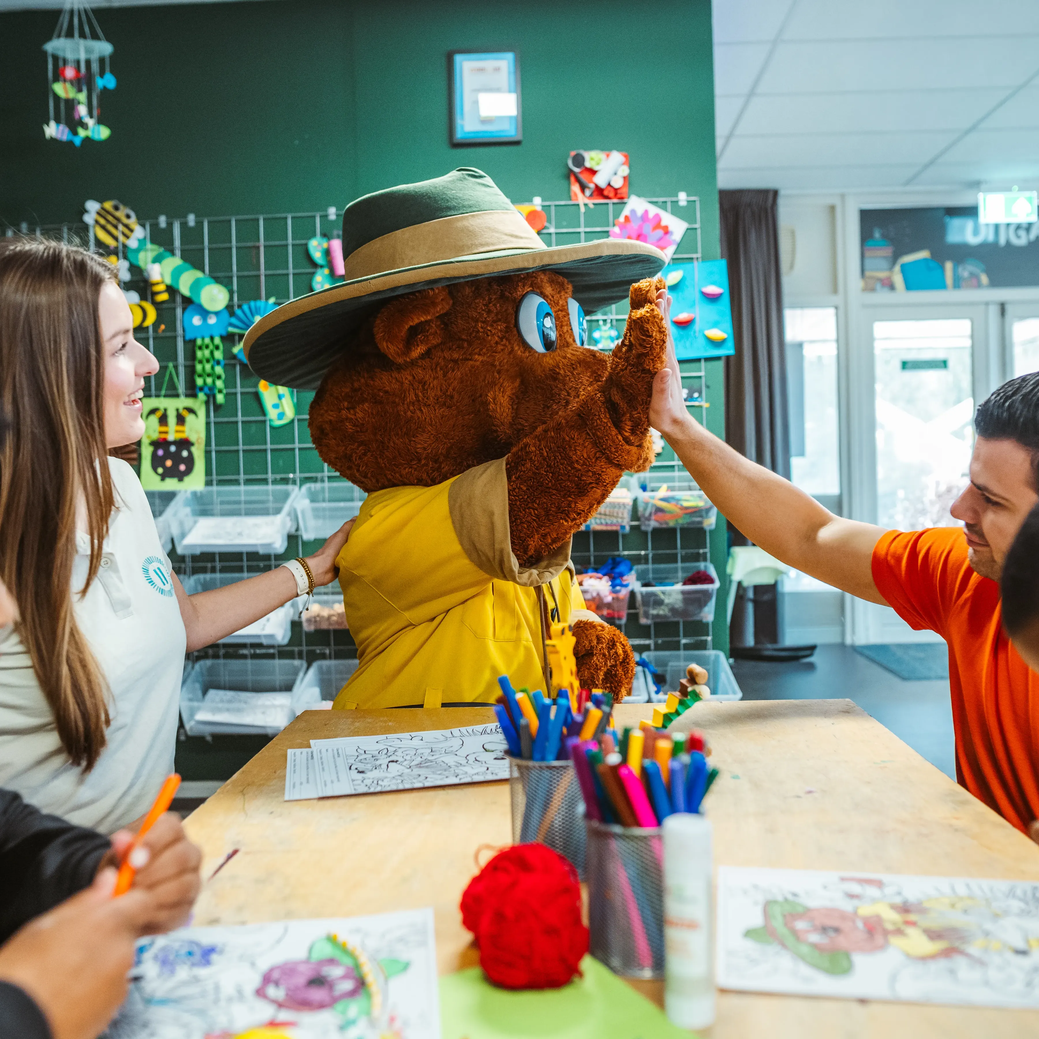 Zwei kleine Jungen sitzen an einem Tisch und malen ein Bollo-Malbild aus. Bollo steht am Ende des Tisches und gibt einem männlichen Landal-Mitarbeiter ein High-Five, während eine Mitarbeiterin Bollo die Hand auf die Schulter legt.