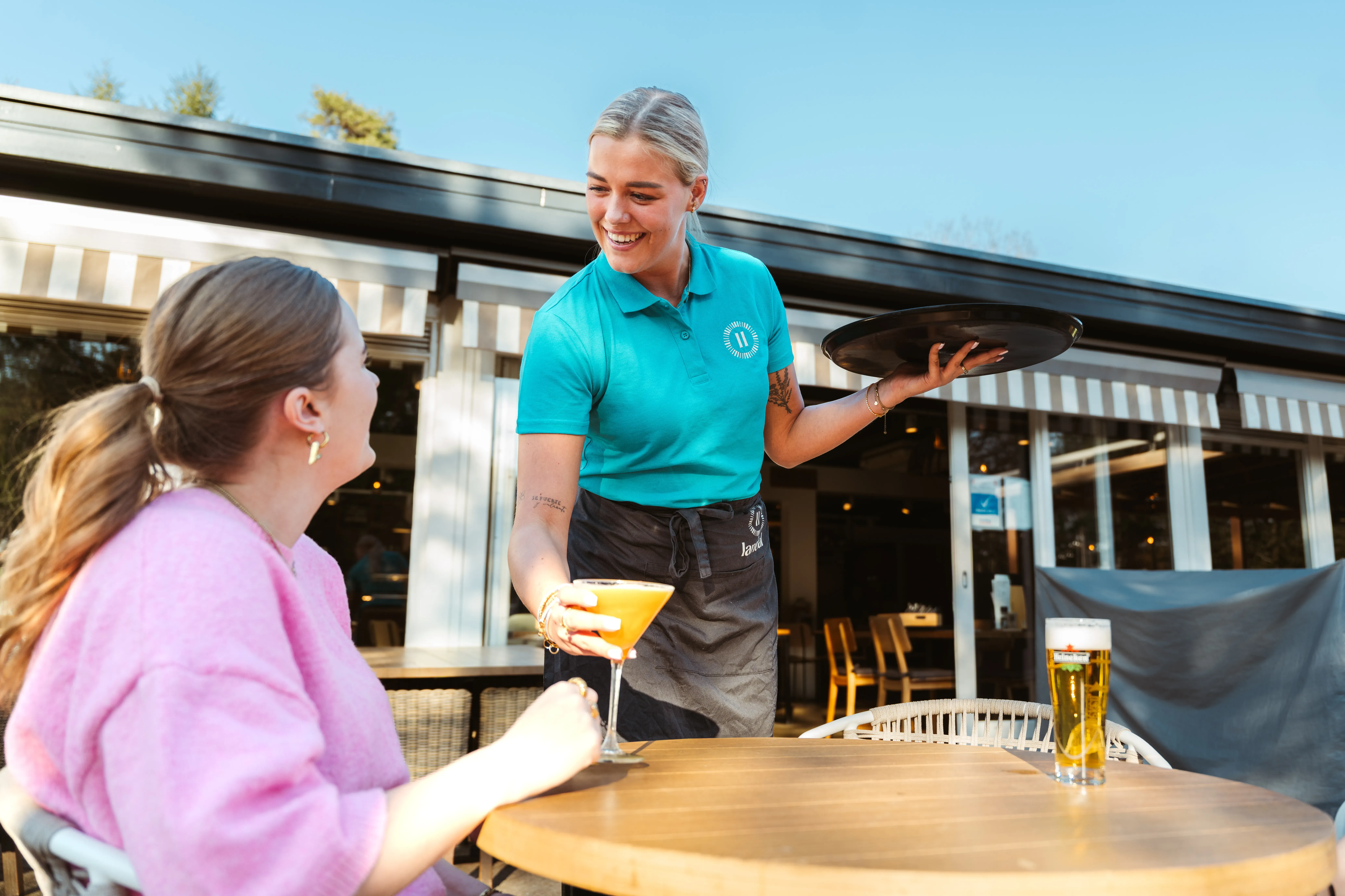 Eine weibliche Angestellte von Landal serviert einem Gast einen Cocktail; beide sehen sich an und lächeln.