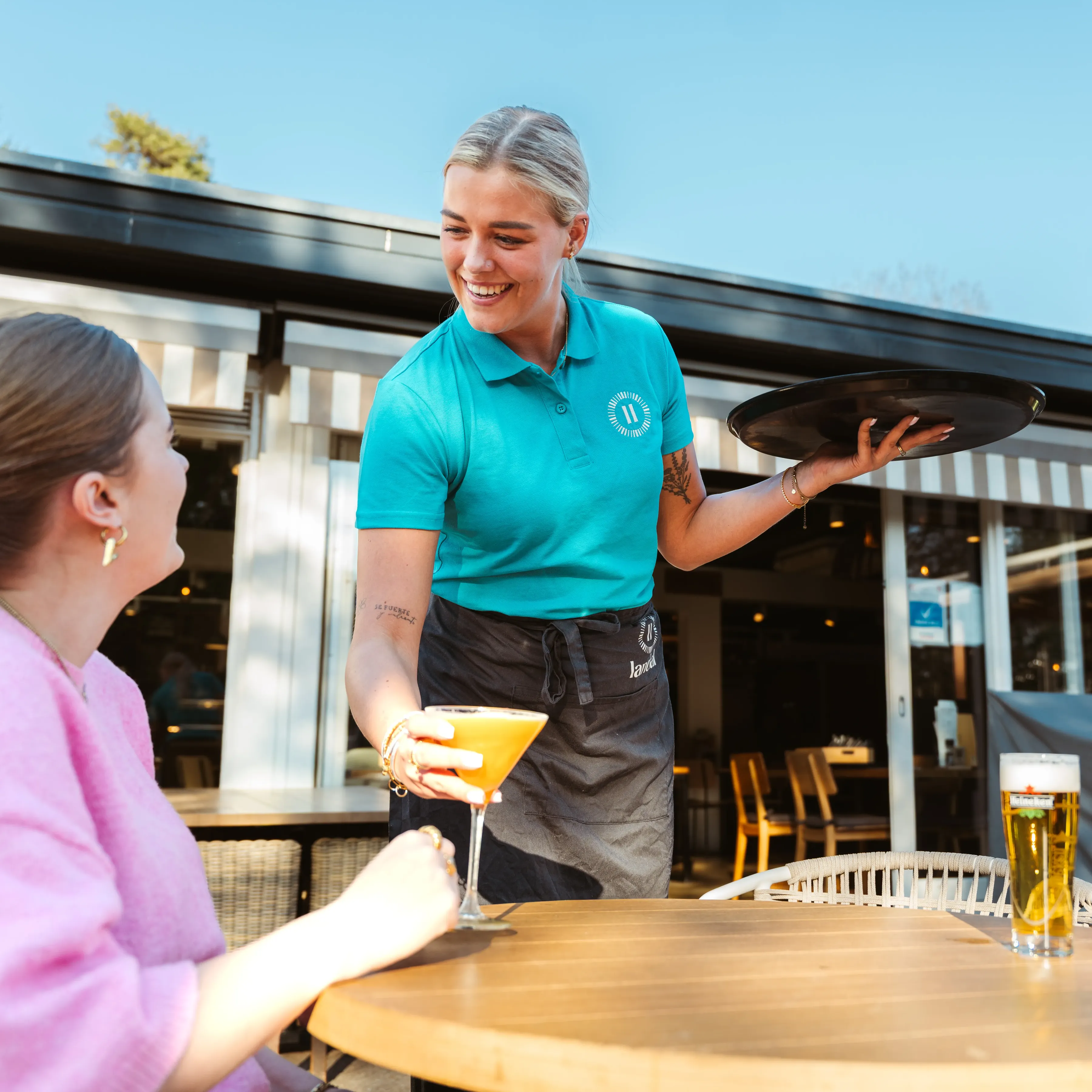 Eine weibliche Angestellte von Landal serviert einem Gast einen Cocktail; beide sehen sich an und lächeln.