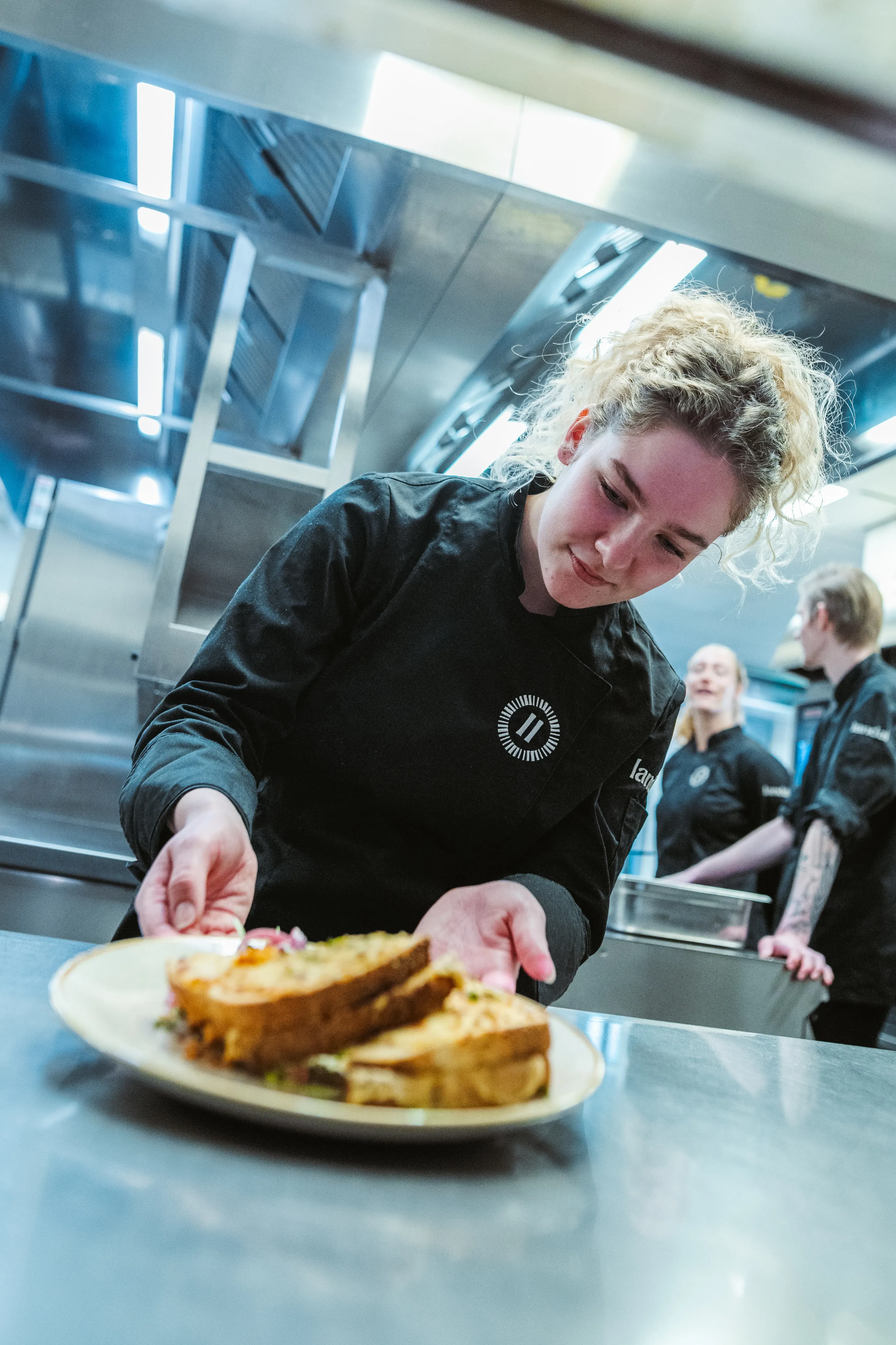 Een jonge Landal-medewerkster in zwarte koksbuis plaatst geconcentreerd een bord met een tosti op het aanrechtblad, terwijl twee collega's op de achtergrond in de professionele keuken werken.