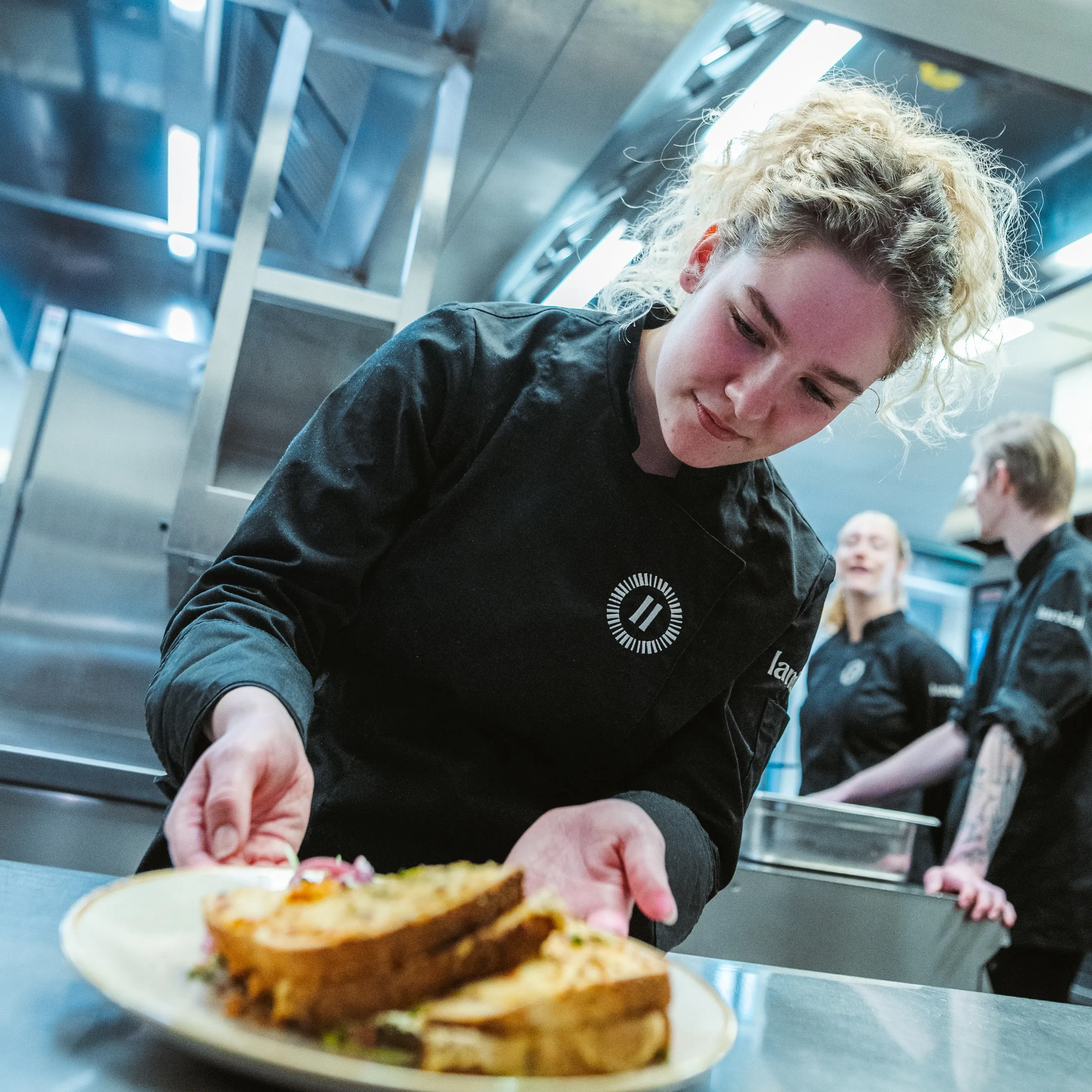 Een jonge Landal-medewerkster in zwarte koksbuis plaatst geconcentreerd een bord met een tosti op het aanrechtblad, terwijl twee collega's op de achtergrond in de professionele keuken werken.